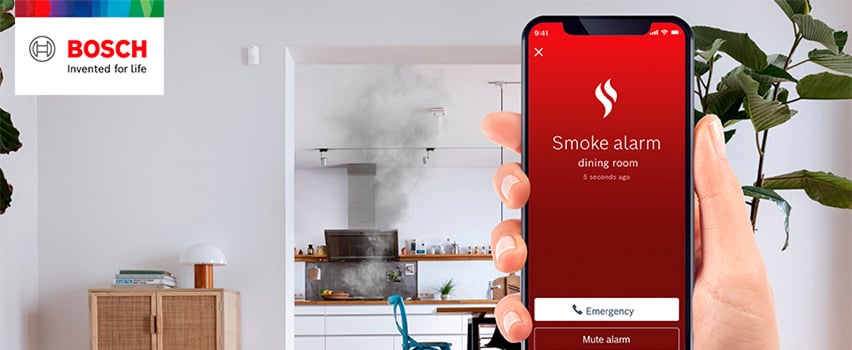Smartphone viser røgalarm-notifikation fra spisestuen, mens røg trænger ud fra køkkenet i baggrunden.