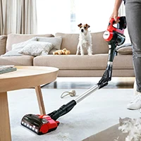 Person støvsuger lyst gulv med rød og sort Bosch trådløs støvsuger med hund på sofa i baggrunden