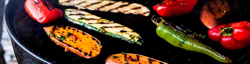 Rød peber, græskar, squash og et halvt løg tilberedes på en lille rund grill på et træbord.