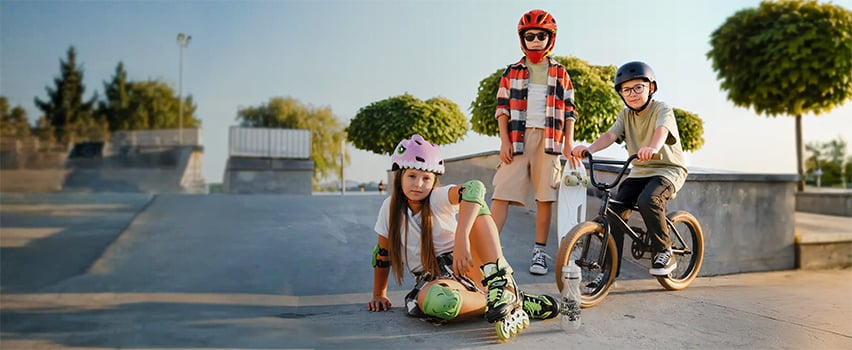 Tre børn med hjelme og beskyttelsesudstyr fra Wozinsky på skateboard, rulleskøjter og BMX-cykel i skatepark