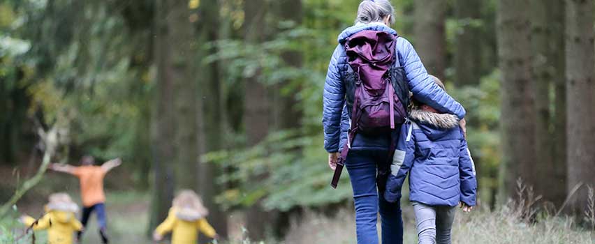 Familie på tur i skov, iført jakker og rygsække, nyder udendørs fællesskab og bevægelse i naturen.