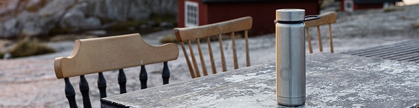 metal termoflaske står på et udendørsbord med røde huse og solopgang i baggrunden.